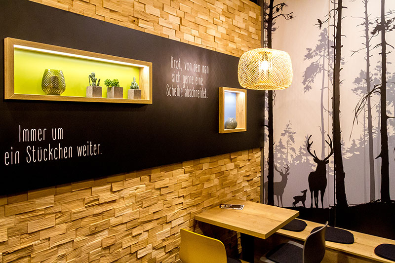 Backsteinwand mit Tafel und eingebauten Regalen. Davor Tisch und Stühle. Die andere Wand ist mit einer Motivtapete im Wald-Stil beklebt