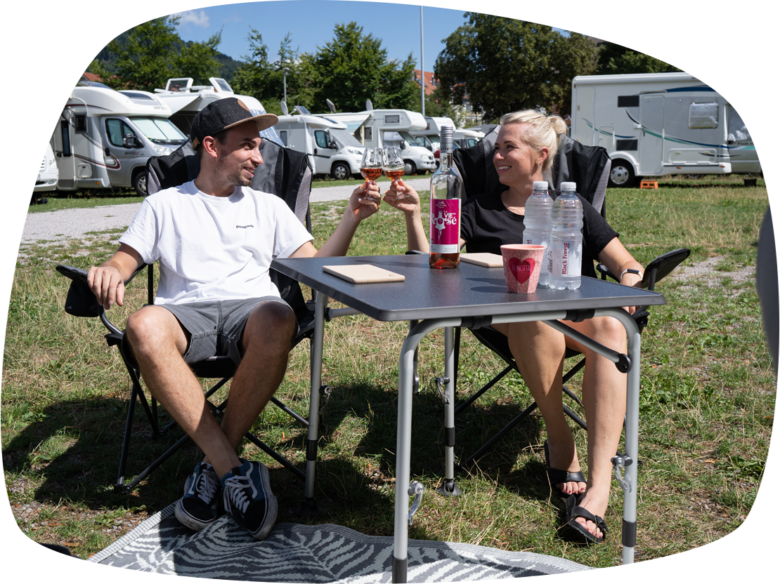 Ein Paar sitzt an einem Campingtisch. Dahinter sind mehrere Wohnmobile zu sehen 
