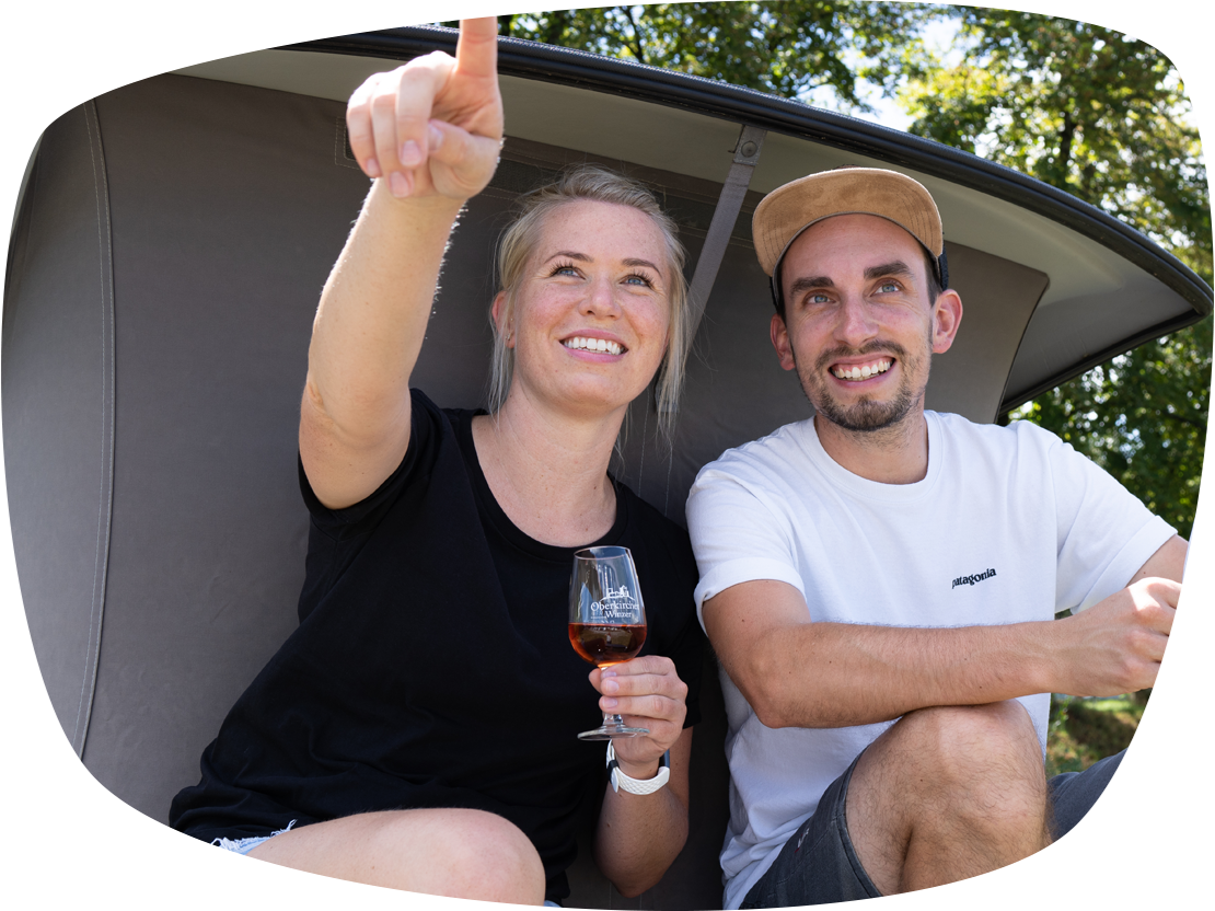 Ein junges Paar vor einem Zelt. Die Frau hat ein Glas Rotwein in der Hand und zeigt auf etwas